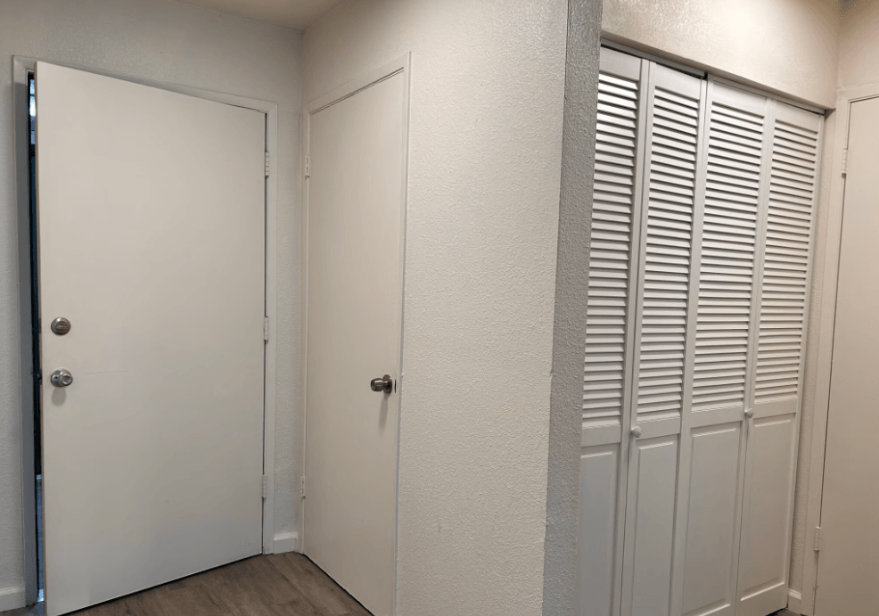 A white door with a silver handle is on the left and a white door with a black handle is in the middle of a hallway with white walls and a white floor.